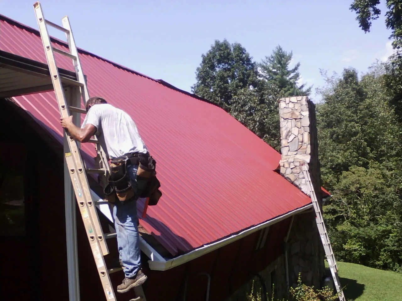 Roofer on a ladder beside a red metal roof and stone chimney — Soffit & Fascia Repair by Roofing Brothers in Cedar Glen